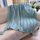 Blue knitted blanket with tassels on a beige couch in a living room setting.