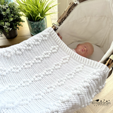 Baby in a crib with a white textured blanket, surrounded by plants and decor.