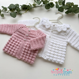 Pink and white crocheted baby cardigans on hangers with greenery in the background, featuring Kerry Jayne Designs logo.