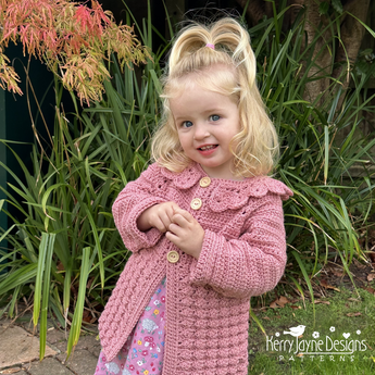 Young girl wearing a pink crochet coat standing outdoors with greenery in the background