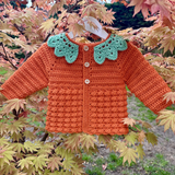 Crocheted orange sweater with green lace collar on a branch with autumn leaves