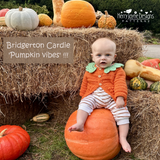 Baby in an orange outfit sitting on a pumpkin with pumpkins and hay bales in the background.