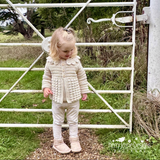 Child wearing a cream crochet jacket standing in front of a white metal gate outdoors.
