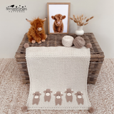 A handmade crocheted blanket with five small cow motifs along the bottom edge, draped over a wicker chest. The blanket is styled with a plush Highland cow toy, a framed photo of a real cow, and yarn balls on top, set on a textured beige carpet.
