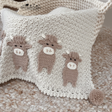 A handmade cream-colored crochet baby blanket featuring three brown crocheted cow appliqués, styled inside a woven cream basket on a textured beige rug. 