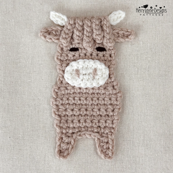 A crocheted plush cow applique in taupe and white, laid flat on a textured beige fabric background. The cow has small black embroidered eyes and a white snout