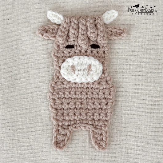 A crocheted plush cow applique in taupe and white, laid flat on a textured beige fabric background. The cow has small black embroidered eyes and a white snout