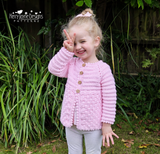 A young girl outdoors wearing a light pink hand-crocheted cardigan with gold buttons, posing in front of green plants. The brand logo 'Kerry Jayne Designs PATTERNS' is visible in the top left corner.