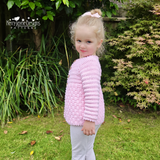 A young girl with blonde hair in a ponytail wearing a light pink crocheted cardigan and light gray pants, standing in a grassy garden with green plants and shrubs.