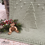A fern green crocheted blanket with a raised Christmas tree design and pom-pom border, draped over a wicker trunk in a styled christmas setting. The scene includes a small teddy bear, a pattern by Kerry Jayne Designs 
