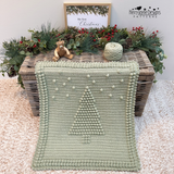 A fern green coloured crocheted blanket with a raised Christmas tree design and pom-pom border, draped over a wicker trunk in a styled christmas setting. The scene includes greenery, pinecones, yarn balls, a small teddy bear, a pattern by Kerry Jayne Designs 