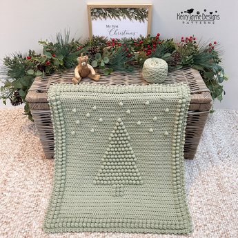 A fern green coloured crocheted blanket with a raised Christmas tree design and pom-pom border, draped over a wicker trunk in a styled christmas setting. The scene includes greenery, pinecones, yarn balls, a small teddy bear, a pattern by Kerry Jayne Designs 