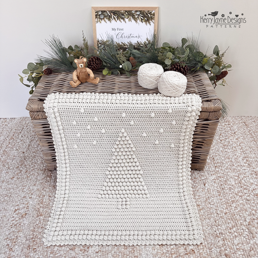 An off-white crocheted blanket with a raised Christmas tree design and pom-pom border, draped over a wicker trunk in a styled christmas setting. The scene includes greenery, pinecones, yarn balls, a small teddy bear, a pattern by Kerry Jayne Designs 