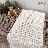 An off-white crocheted blanket with a raised Christmas tree design and pom-pom border, draped over a wicker trunk in a styled christmas setting. The scene includes greenery, pinecones, yarn balls, a small teddy bear, a pattern by Kerry Jayne Designs 
