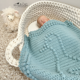 A newborn baby is sleeping in a white rope basket, wrapped in a light blue textured knit blanket. A white plush sheep toy sits on the beige carpet beside the basket.
