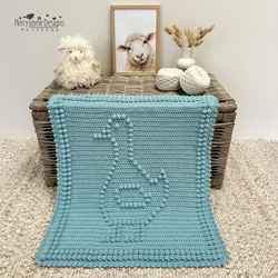 A crocheted baby blanket in light blue with a raised goose design and pom-pom border, draped over a wicker chest in a styled nursery setting. A plush sheep toy, a framed sheep portrait, yarn balls, and a small vase of dried grasses sit on the chest.