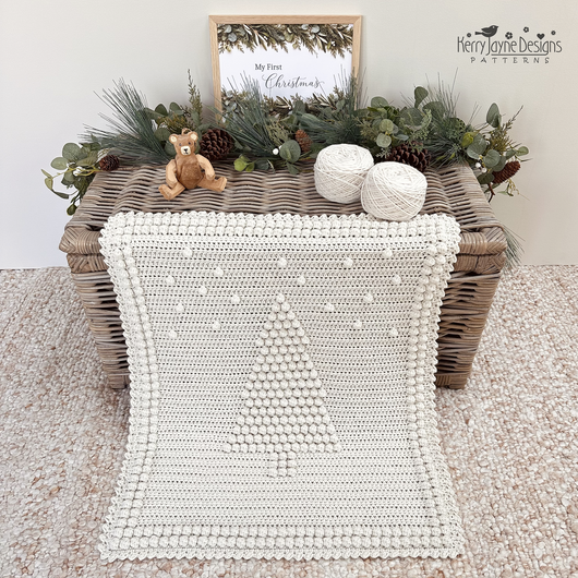 An off-white crocheted blanket with a raised Christmas tree design and pom-pom border, draped over a wicker trunk in a styled christmas setting. The scene includes greenery, pinecones, yarn balls, a small teddy bear, a pattern by Kerry Jayne Designs 