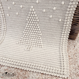 An off-white crocheted blanket with a raised Christmas tree design and pom-pom border, draped over a wicker trunk in a styled christmas setting. A crochet pattern by Kerry Jayne Designs 