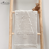 An off-white crocheted blanket with a raised Christmas tree design and pom-pom border, draped over a styled ladder. The scene includes greenery, pinecones, yarn balls, a small teddy bear, a pattern by Kerry Jayne Designs 