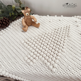 An off-white crocheted blanket with a raised Christmas tree design and pom-pom border, draped over a wicker trunk in a styled christmas setting. The scene includes greenery, pinecones, yarn balls, a small teddy bear, a pattern by Kerry Jayne Designs 