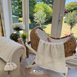 Wicker chair with a textured blanket draped over it, surrounded by plants and a teddy bear in a sunroom.