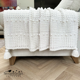 White crocheted blanket with tassels draped over a sofa on a wooden floor.