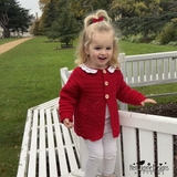 Child wearing a red crocheted cardigan standing on a white bench outdoors.