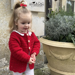 Child wearing a red crocheted cardigan standing next to a large plant pot.