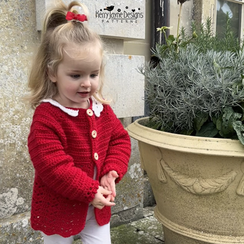 Child wearing a red crocheted cardigan standing next to a large plant pot.