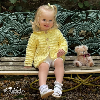 Child wearing a yellow crocheted cardigan sitting on a bench with a teddy bear.