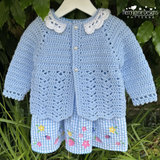 Crocheted baby outfit with blue jacket and shorts on a hanger against a green leafy background.