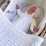 Newborn baby in a white crocheted outfit and bonnet lying in a crib with a textured blanket.