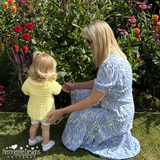 Woman in a blue dress kneeling on grass next to a child in a yellow outfit, surrounded by flowers.