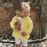 Child wearing a yellow crochet sweater holding a teddy bear in front of a stone wall.