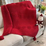 Red knitted blanket with tassels draped over a couch, featuring a small Santa Claus toy.