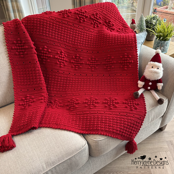 Red knitted blanket with tassels draped over a couch, featuring a small Santa Claus toy.