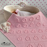 Pink crocheted blanket with a baby in a basket and a stuffed rabbit toy on a heart-patterned background.